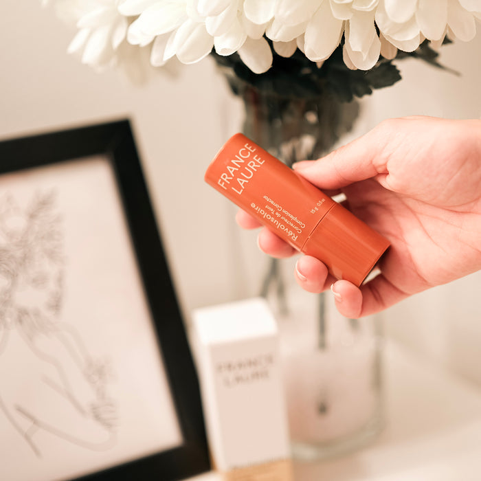 Hand holding France Laure Révolusolaire complexion corrector stick, ideal for all skin types, with white flowers and framed art in soft background.