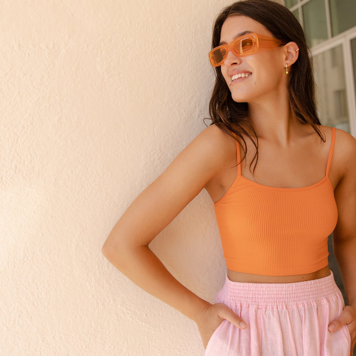 Woman wearing orange square sunglasses and matching ribbed crop top, smiling against a textured beige wall
