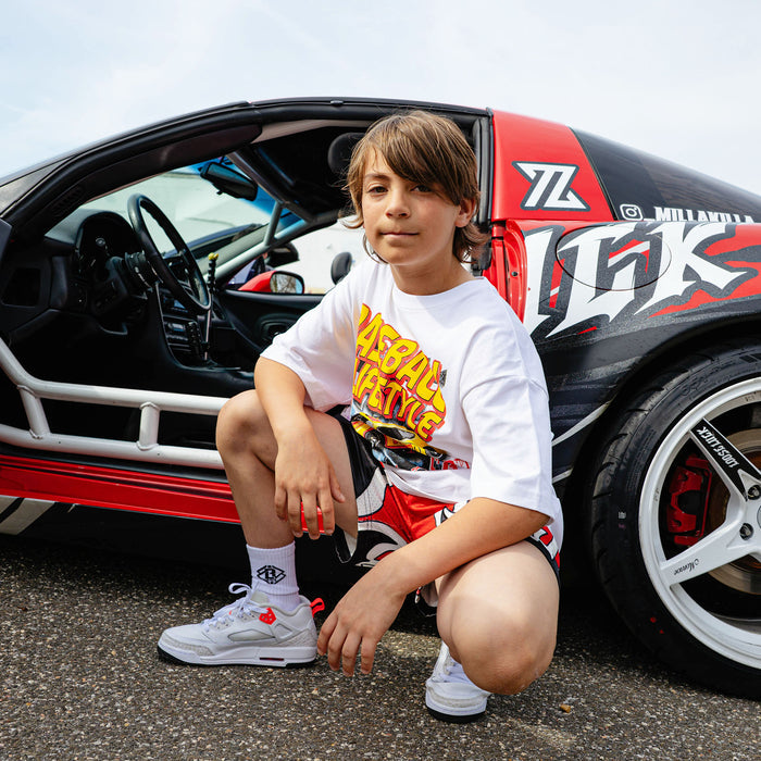 Young boy crouching beside open-door wrapped drift car, wearing white Diamond Drift tee with BL101 race car graphic and red flame shorts.