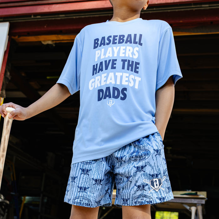 young boy wearing the columbia blue youth Greatest Dad Athletic Tee paired with Father & Son Bat Company all-over print shorts while holding a baseball bat.