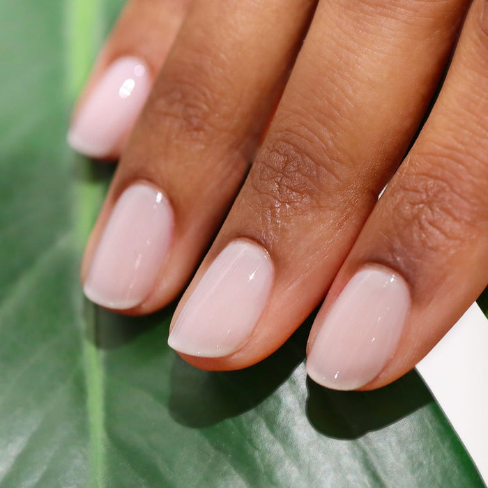 A close-up of a hand with neatly manicured nails painted in Manucurist's Milky White—a sheer, glossy polish—resting on a green leaf background.