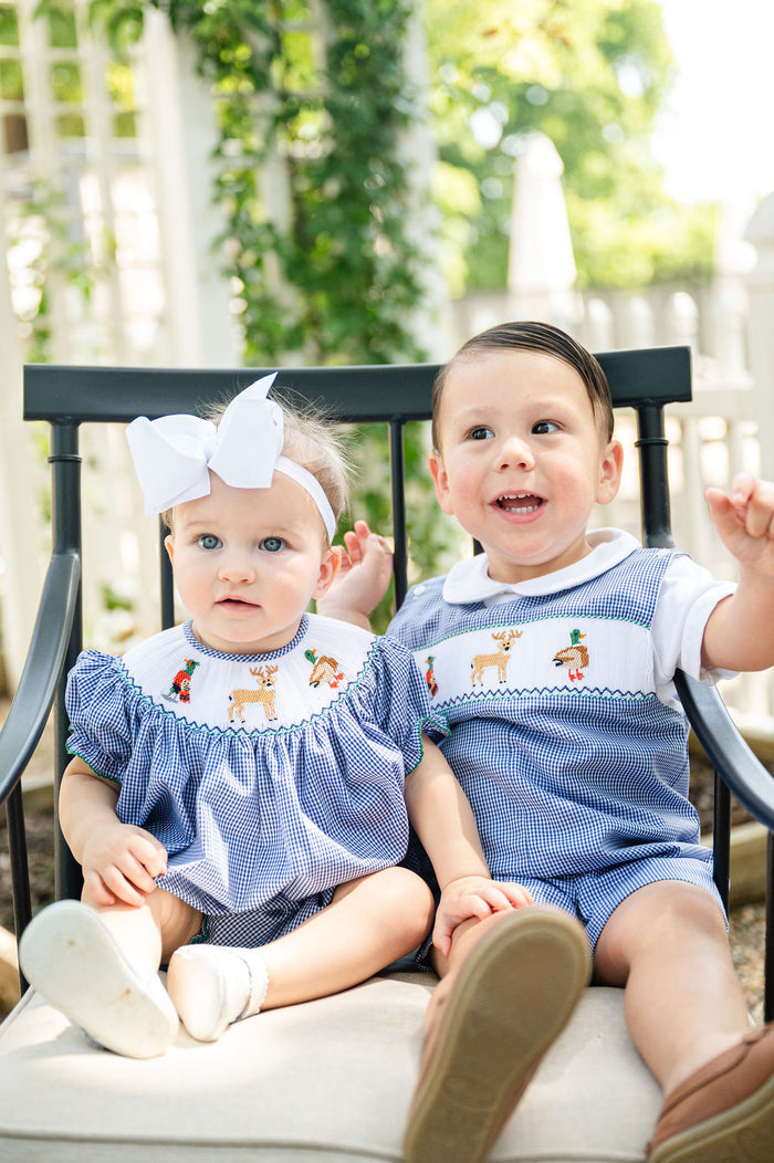 Baby girl in Smocked Hunting Season Bubble by Ruth and Ralph seated outdoors with toddler in matching smocked hunting outfit
