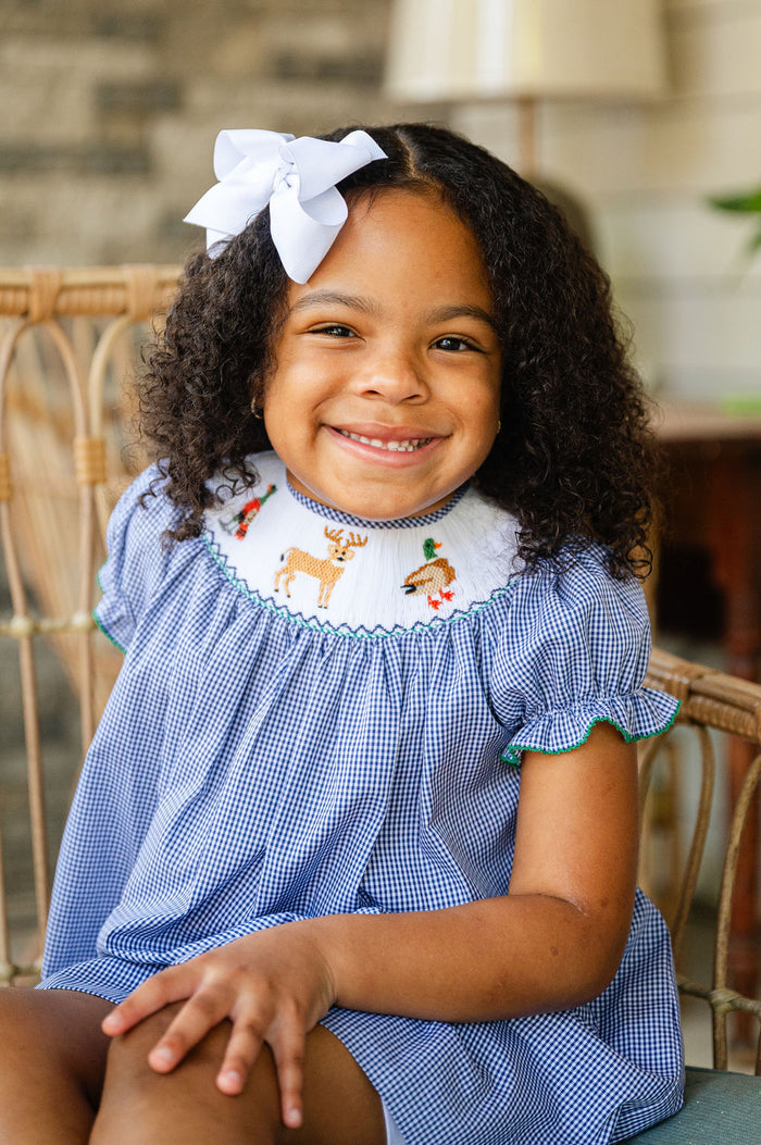 Girl sitting on chair wearing Smocked Hunting Season Dress by Ruth and Ralph, smiling with white hair bow