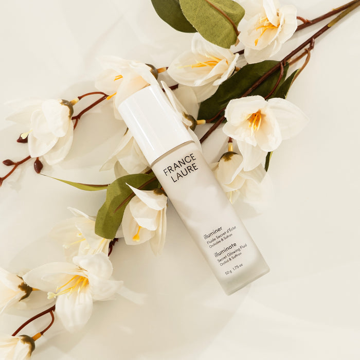France Laure Illuminate Secret Glowing Fluid with orchid & saffron, nestled among white flowers and green leaves on soft fabric