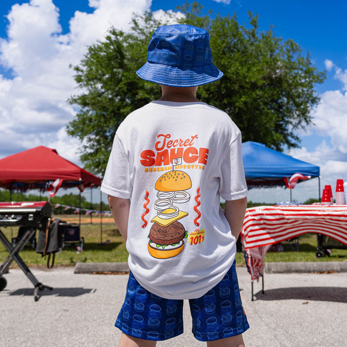 Person wearing a white t-shirt with a graphic design and text, standing outdoors with tents and tables in the background.