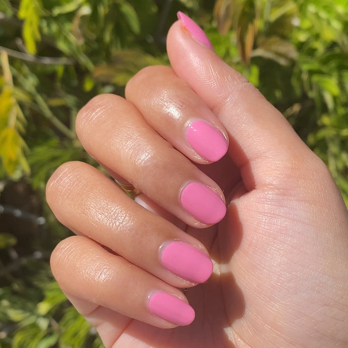 Hand with nails painted in bright pink Taffy gel polish shown outdoors with natural green leafy background