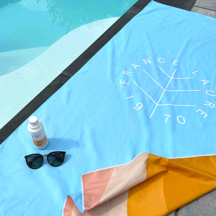 Blue France Laure beach towel with geometric logo, sunscreen SPF 30, and sunglasses by poolside for summer skincare routine