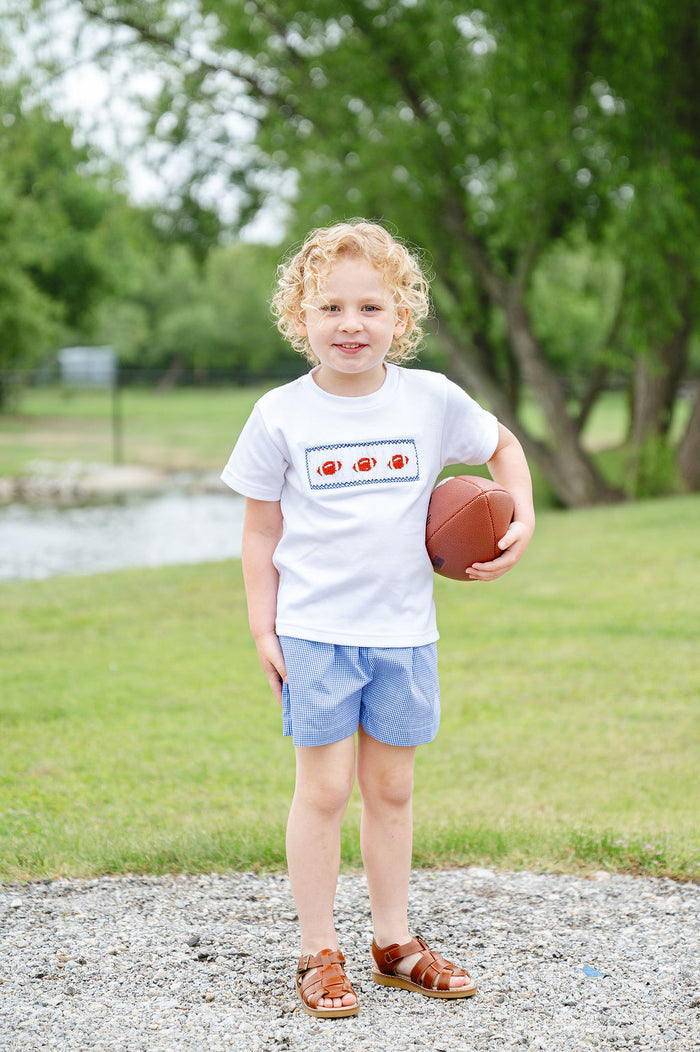 Toddler boy wearing a white smocked football shirt with three hand-smocked footballs and blue gingham shorts, holding a football