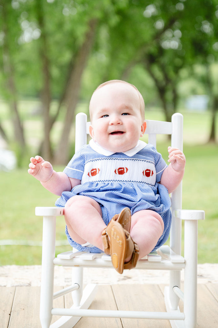 Baby boy wearing a blue gingham smocked football bubble with three hand-smocked footballs and a collared neckline, sitting in a white rocking chair