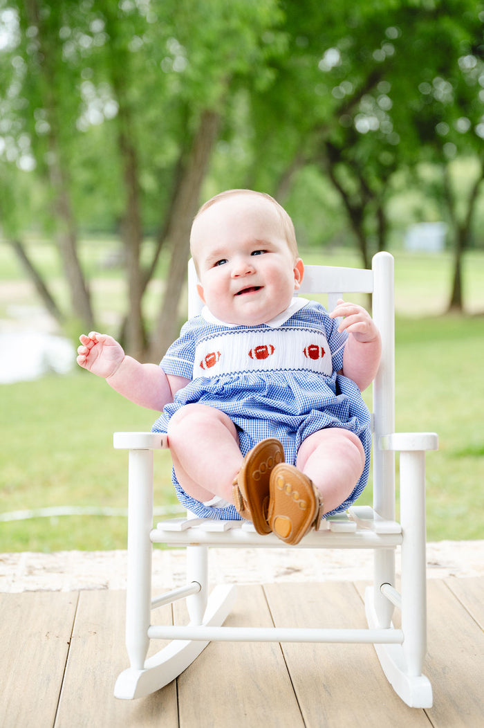 Smiling baby boy in a blue gingham bubble with a hand-smocked football panel, seated outdoors