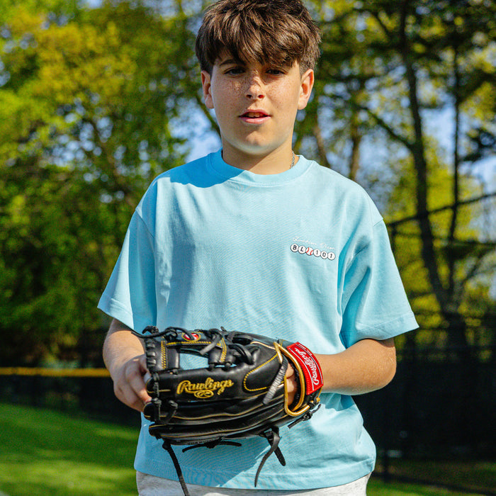 Youth player holding a baseball glove on a field, wearing the light blue Jackson Olson x BL101 BSBL Era relaxed tee with chest logo