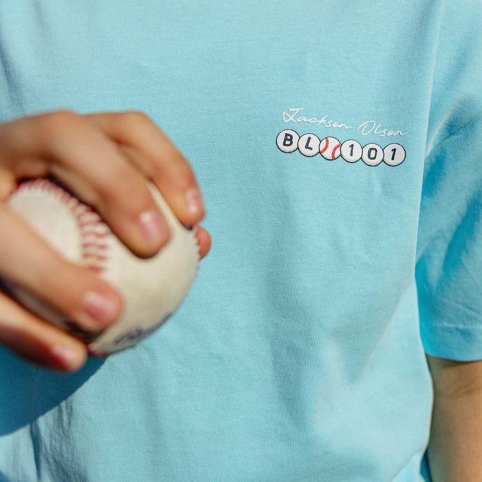 Close-up of the chest graphic on the youth light blue BSBL Era tee, showing the "Jackson Olson" script above the BL101 baseball bead logo
