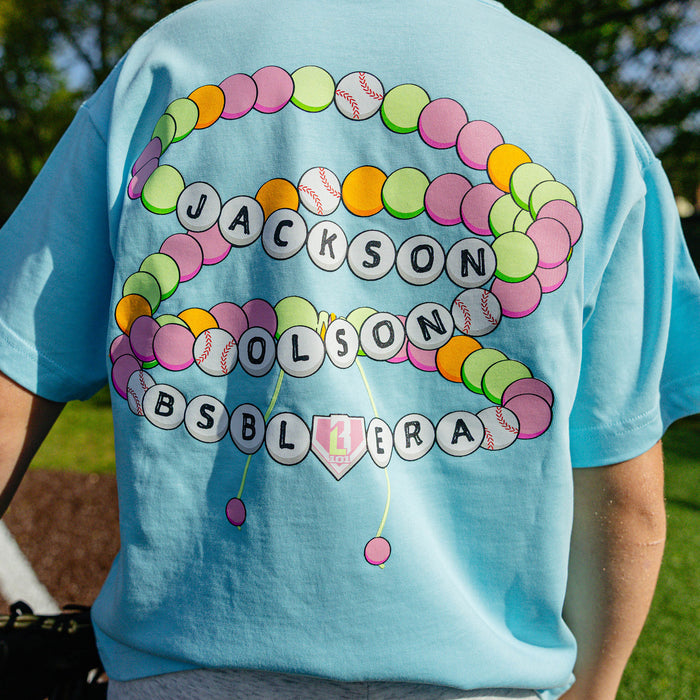 Back view of the youth light blue BSBL Era tee showing the colorful friendship bracelet graphic with "Jackson Olson BSBL Era" lettering and baseball bead details