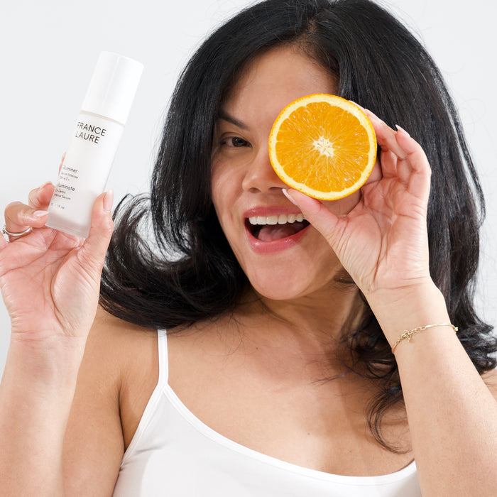 Woman holding France Laure Illuminate C+ Detox Intense Serum and orange slice, showcasing bright skin and vitamin C benefits for radiant complexion.