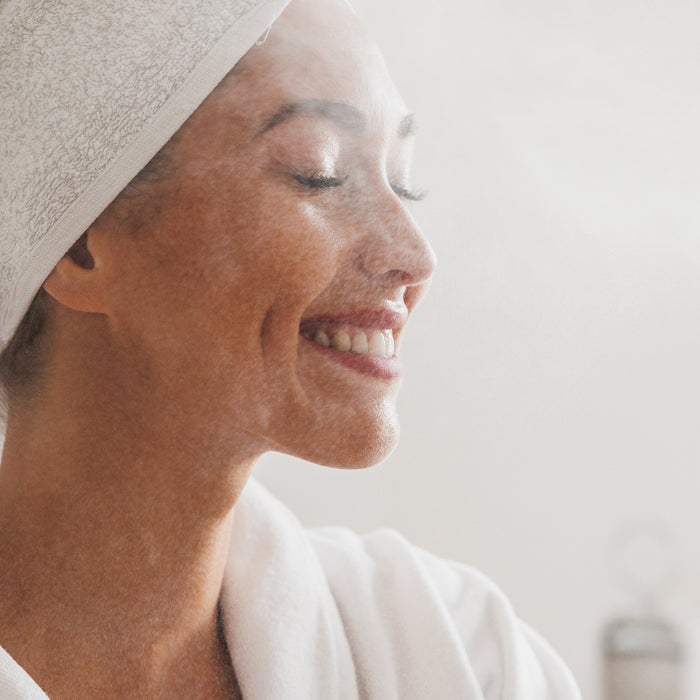Woman smiling with towel-wrapped hair, enjoying gentle facial steam for radiant skin care routine