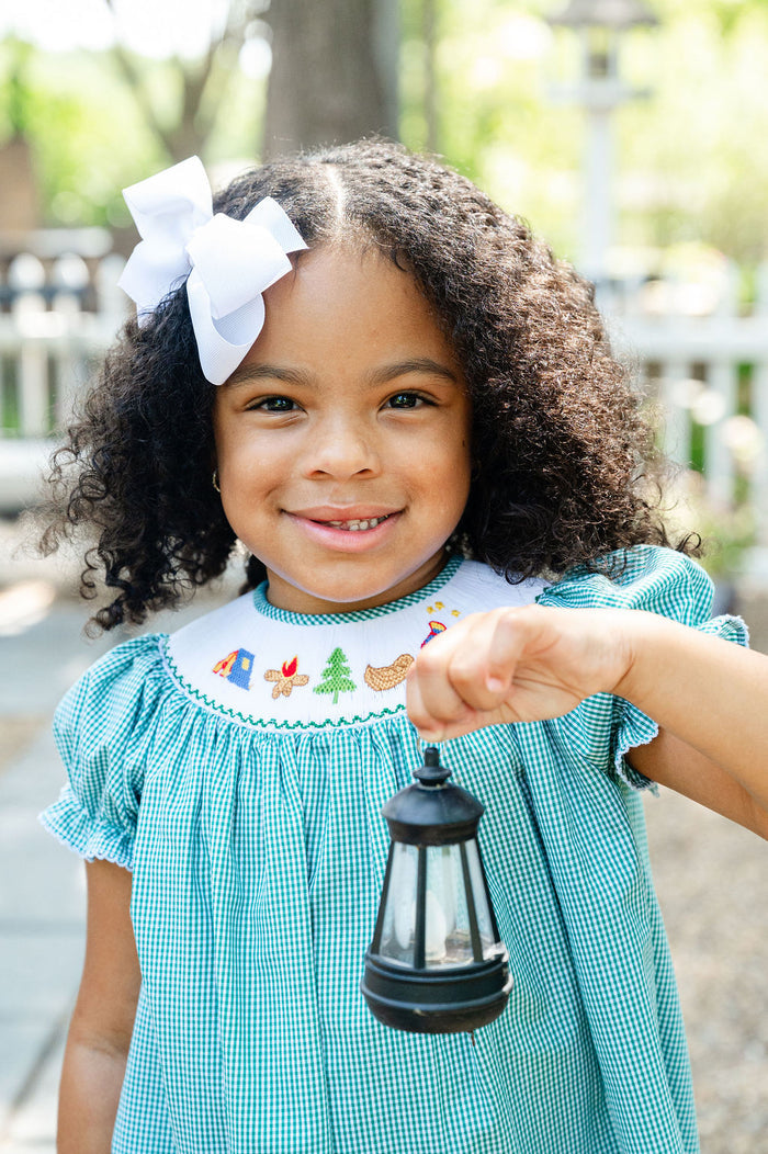 Girl smiling in Smocked Camping Dress by Ruth and Ralph, teal gingham, holding toy lantern outdoors