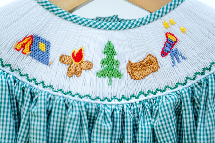Close-up of smocked camping embroidery detail on Ruth and Ralph teal gingham dress, featuring tent, campfire, and tree