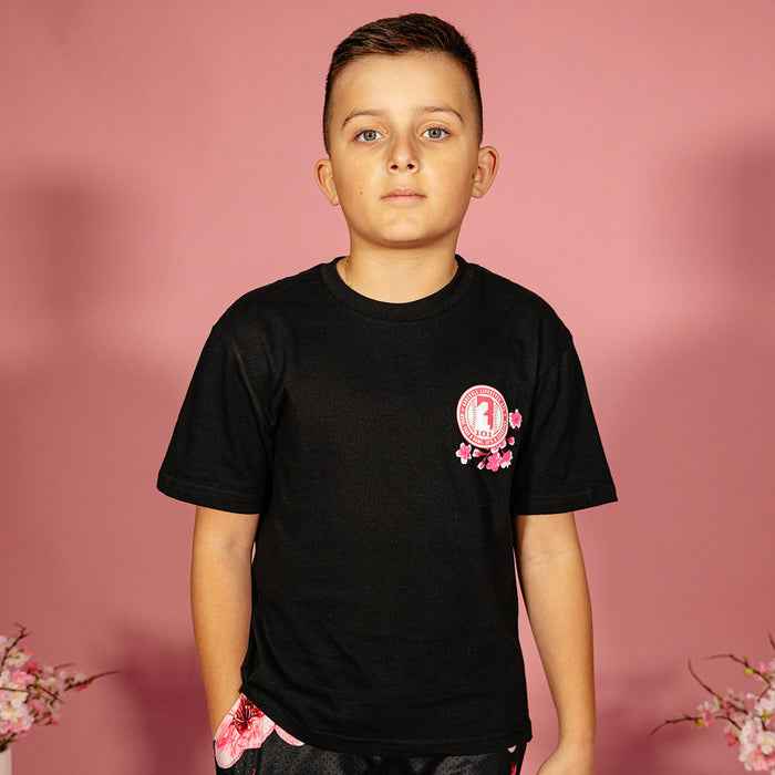 Young boy wearing a black BL101 cherry blossom tee with pink seal logo on the left chest against a pink background, front view