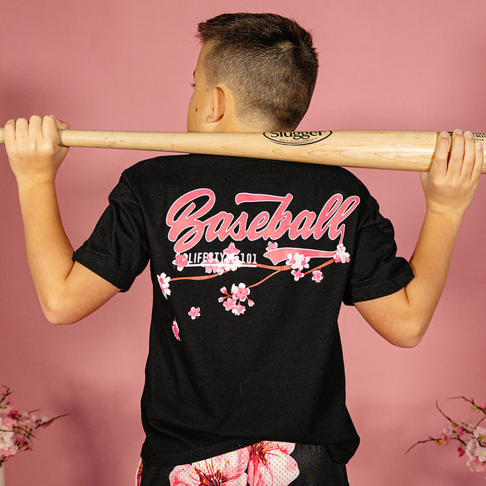 Young boy holding a wooden bat across his shoulders showing the back of a black BL101 tee with pink "Baseball" script and cherry blossom graphic against a pink background