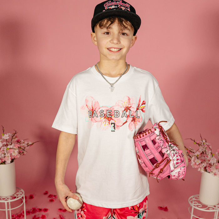 Young boy wearing a white BL101 cherry blossom Baseball tee, black cherry blossom snapback, and red floral shorts, holding a baseball and pink floral glove against a pink background