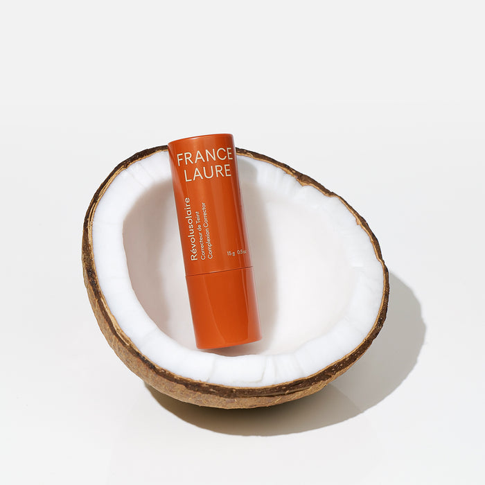 France Laure Révolusolaire complexion corrector stick in orange tube, nestled in a coconut half on white background