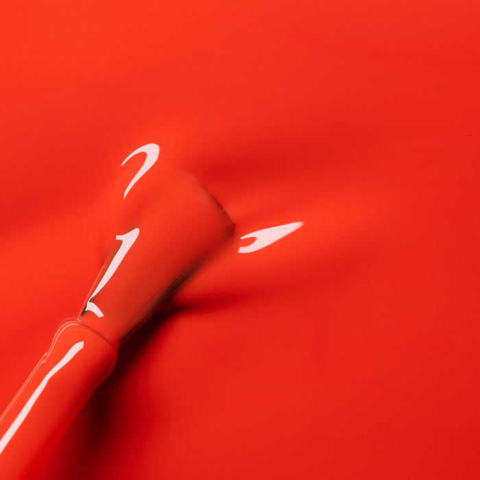 A close-up of a Manucurist Coral Reef brush spreading vibrant orange-red polish on a smooth surface, revealing glossy reflections in the wet finish.