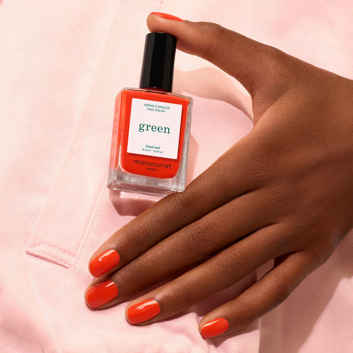A hand with Coral Reef nail polish by Manucurist holds a matching bottle of the polish against a light pink fabric background.