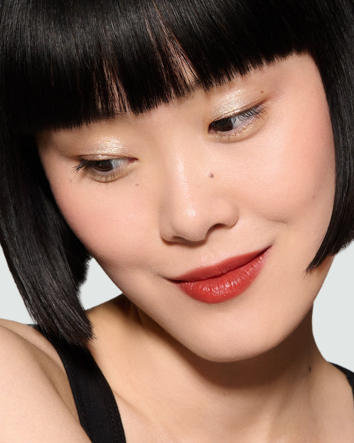 Close-up of a model with black hair and red Lili Rouge Experience Lipstick and Desert Gleam Eyeshadow on, against a plain background