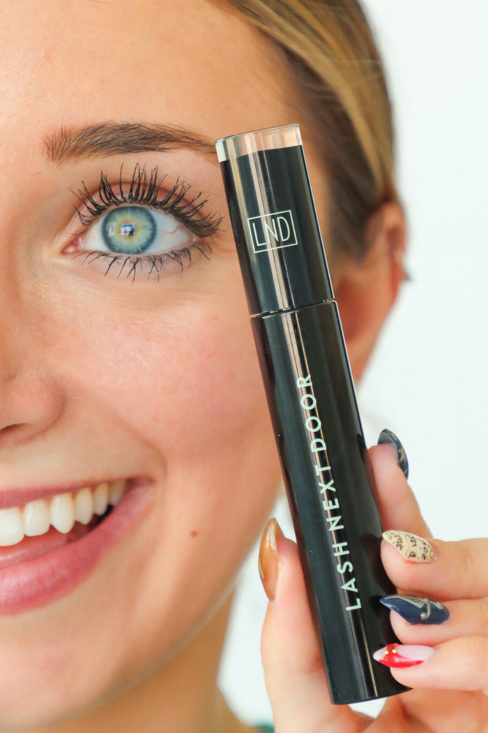 Woman holding a black mascara tube labeled 'Lash Next Door' with a blurred background