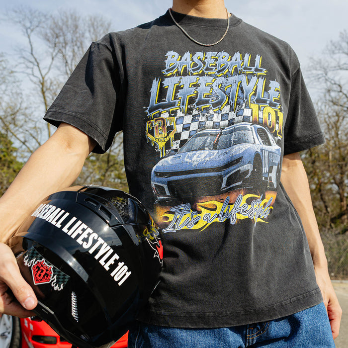 Close-up of washed black youth Fast Track Fielder tee showing vintage race car, checkered flag, and "It's a Lifestyle" flame script graphic. Model holding baseball bat beside silver car.