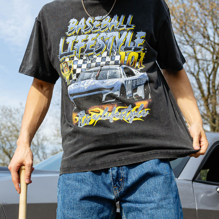 Cropped front view of washed black youth tee with full race car and flame graphic, model holding baseball bat beside silver car.