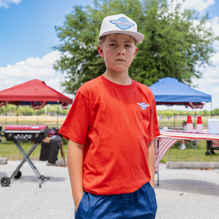 Front-facing youth male model wearing red Fastball Flight tee with gray Fastball Flight snapback at outdoor baseball event