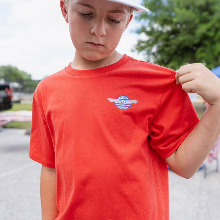 Close-up of youth male model in red Fastball Flight tee pulling shirt to show Air Show Division badge on chest