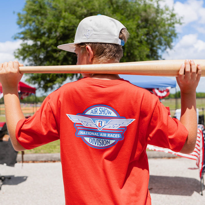 Back view of youth male model holding baseball bat across shoulders showing Air Show National Air Races Division badge on red tee at outdoor event