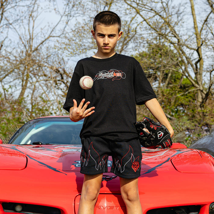 oung boy standing on red Corvette hood tossing a baseball, wearing black Flames Stitched tee with BL101 graphic and black flame shorts, holding baseball glove.