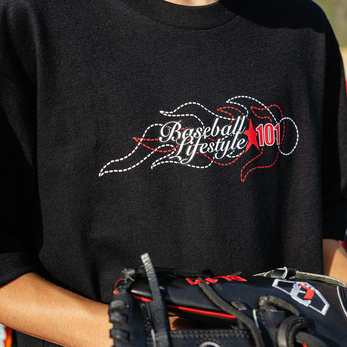 Close-up of stitched Baseball Lifestyle 101 flame chest graphic on black youth tee, baseball glove visible at bottom.