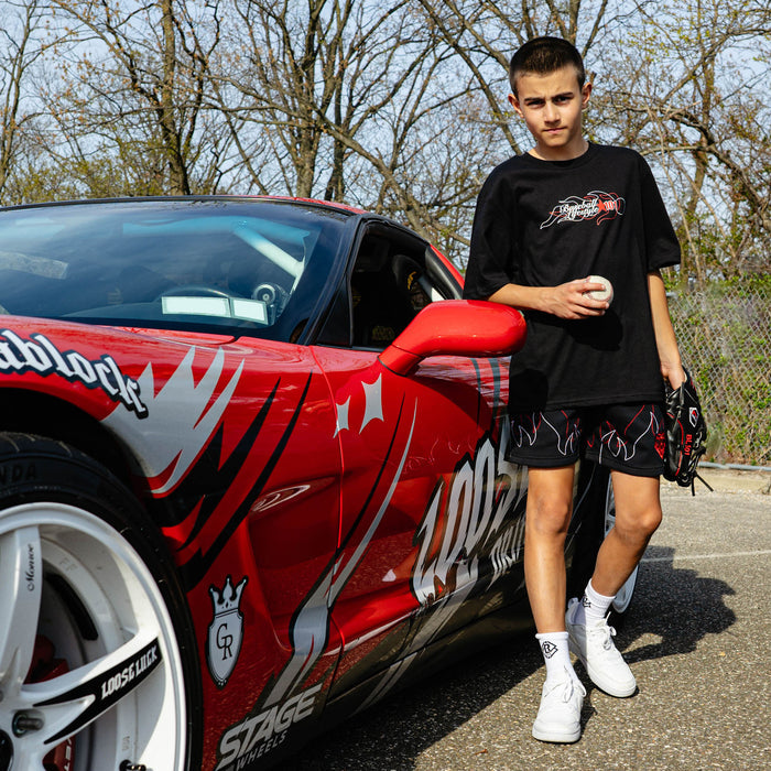 Young boy leaning against wrapped red sports car holding baseball and glove, wearing black Flames Stitched tee and black flame shorts.