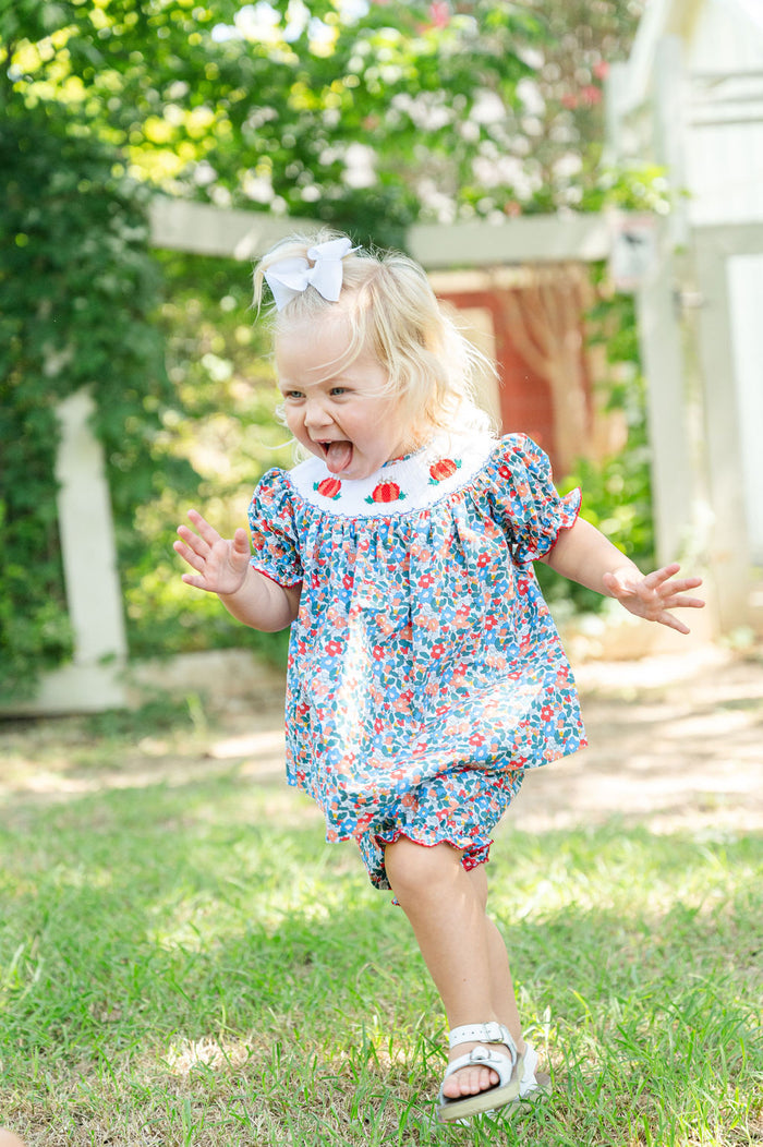 Baby girl running and laughing in Smocked Floral Pumpkin Bloomer Set by Ruth and Ralph, blue floral print with white bow, outdoors