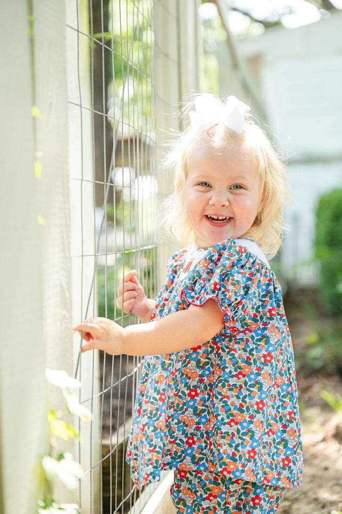 Baby girl smiling in doorway wearing Smocked Floral Pumpkin Bloomer Set by Ruth and Ralph, blue floral print with white bow