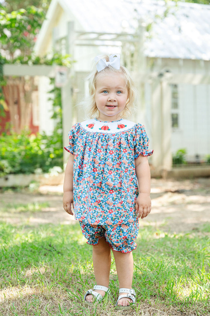 Baby girl standing outdoors wearing Smocked Floral Pumpkin Bloomer Set by Ruth and Ralph, blue floral print