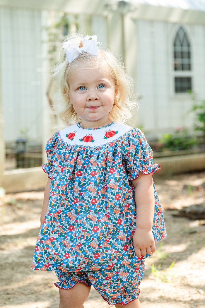 Baby girl standing outdoors wearing Smocked Floral Pumpkin Bloomer Set by Ruth and Ralph, blue floral print with white bow