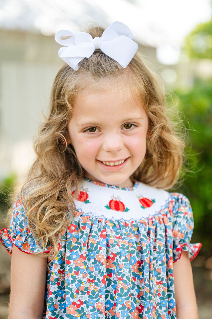 Girl smiling in Smocked Floral Pumpkin Dress by Ruth and Ralph, blue floral print with white bow, outdoors