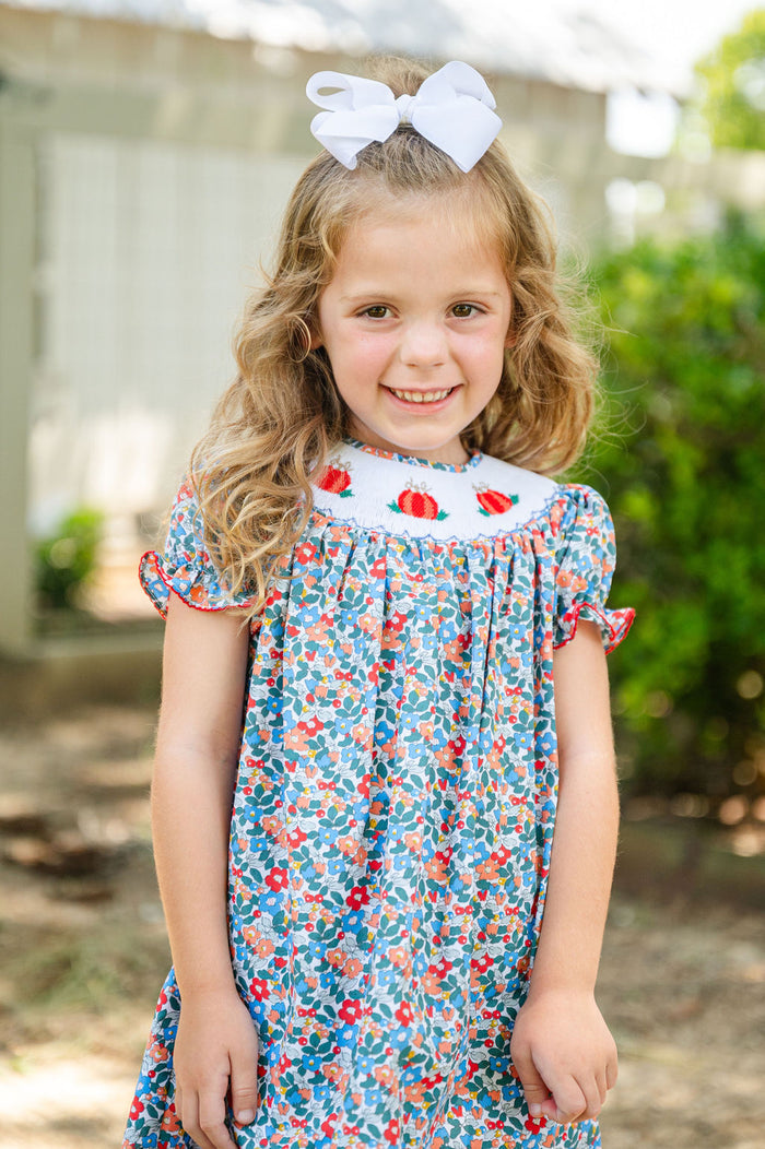 Girl standing outdoors in Ruth and Ralph Smocked Floral Pumpkin Dress, blue floral print with white bow