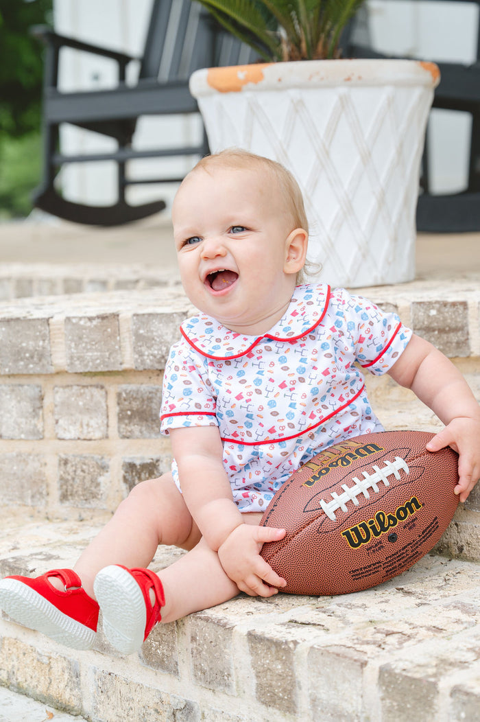 Baby wearing a white Pima cotton football bubble with game day print, red piping, and rounded collar, holding a Wilson football