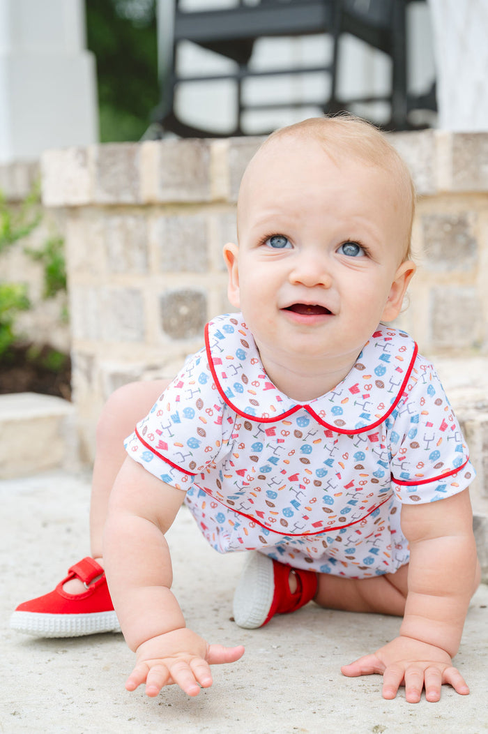 Smiling baby in a white and red game day bubble with football and tailgate motif print, sitting on a brick step