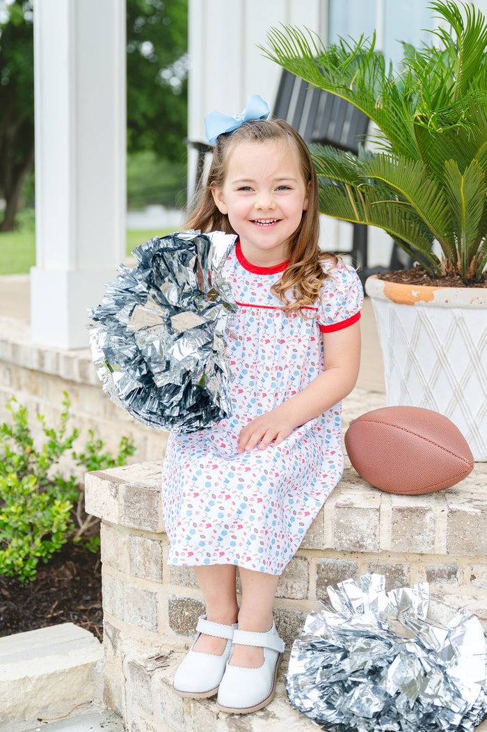 Toddler girl wearing a white Pima cotton football dress with game day print and red trim, holding silver pom-poms