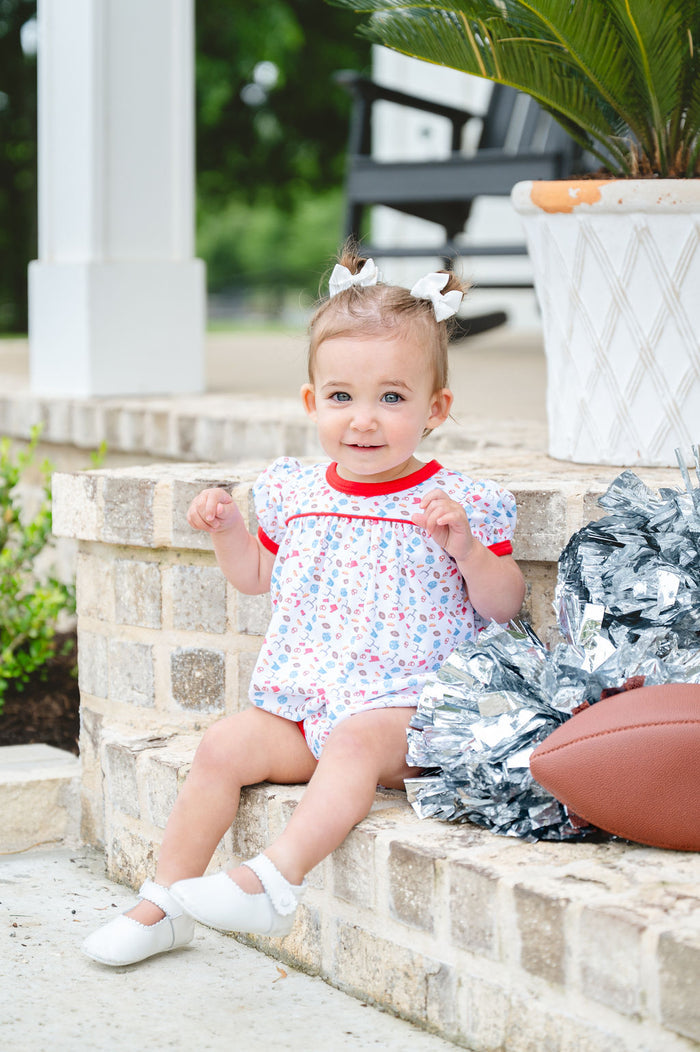 Baby girl wearing a white Pima cotton football bubble with game day print and red trim, with white bows and silver pom-poms