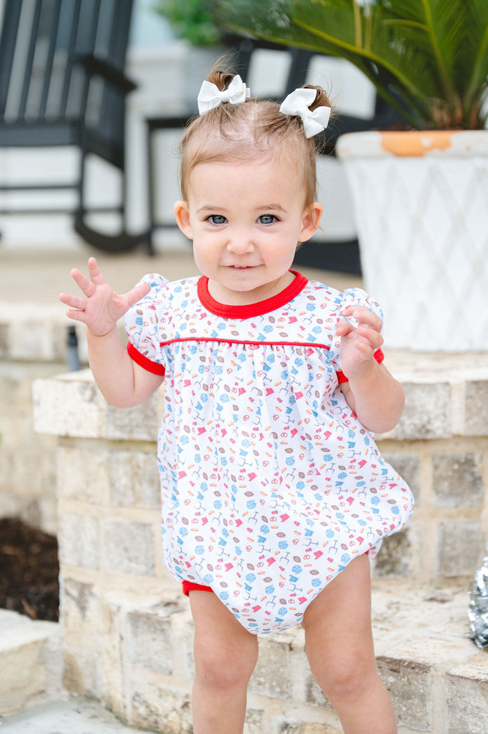 Smiling toddler girl in a white and red game day bubble with football and tailgate motif print, sitting on a brick step