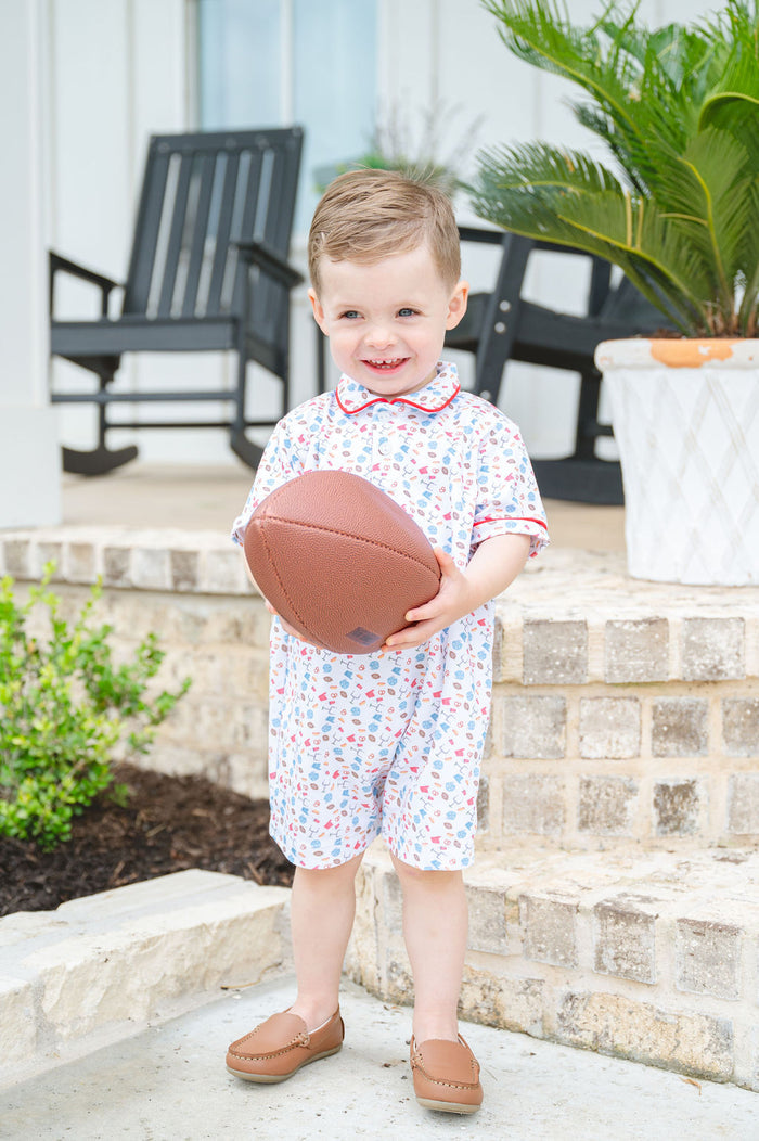 Toddler boy wearing a white Pima cotton football romper with game day print, red piping, and polo collar, holding a football
