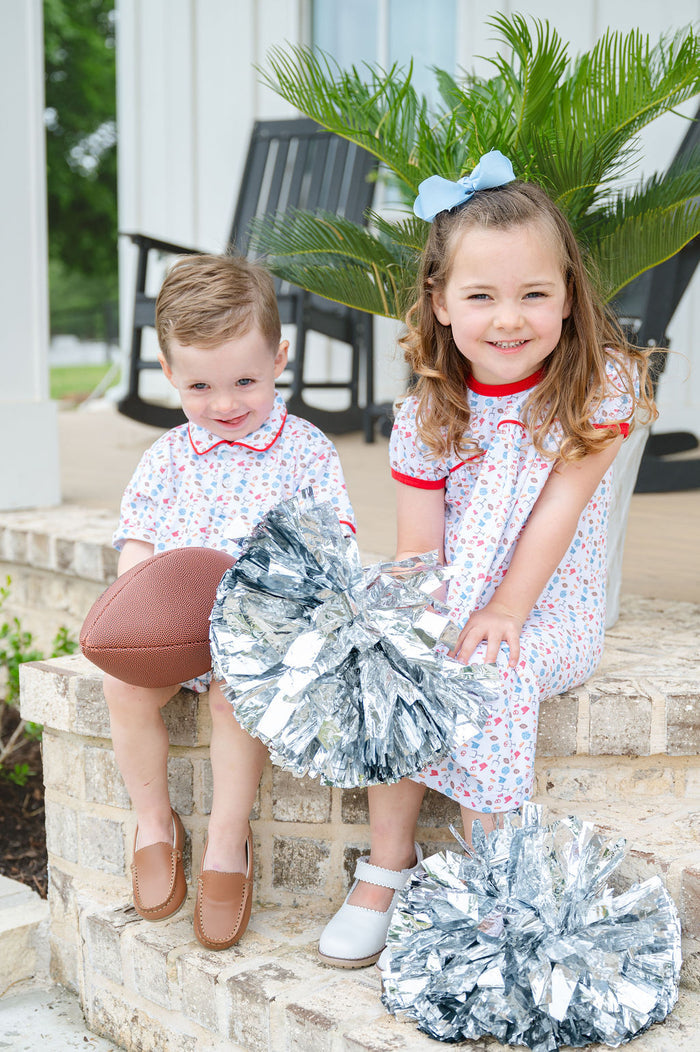 Little boy in a white and red game day romper with football print, standing on a brick porch holding a football